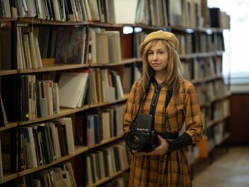 Kobieta w berecie stojąca w bibliotece z aparatem fotograficznym w dłoniach